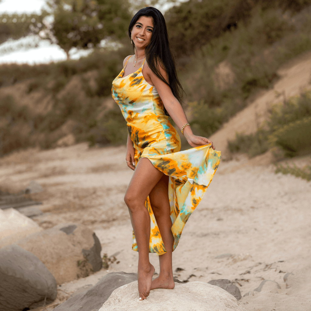 Model wearing a vibrant yellow and blue tie-dye dress, posing on a sandy beach.